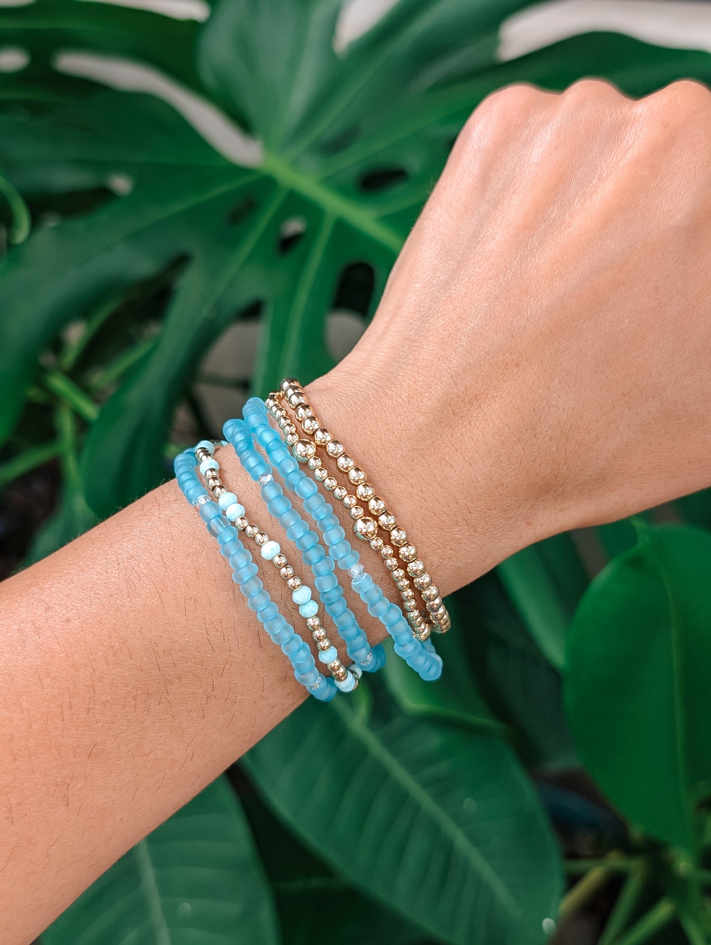Wrist with blue beaded bracelets against a green leafy background